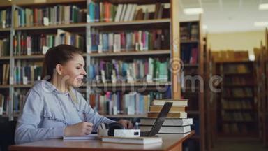 年轻美丽的微笑学生女孩坐在大图书馆的桌子上，有一堆书。 她在写